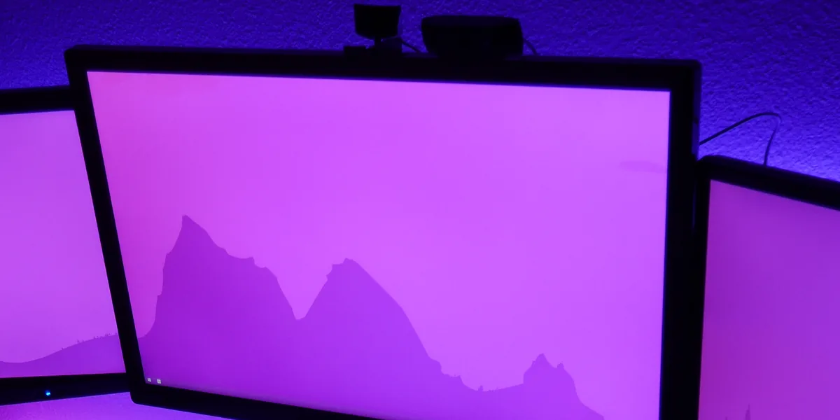 Großer Monitor zwischen den Rändern zweier weiterer Bildschirme, über alle läuft dasselbe violette Hintergrundbild mit dem Umriss einer Gebirgskette, oben auf dem Rahmen zwei kleine Geräte